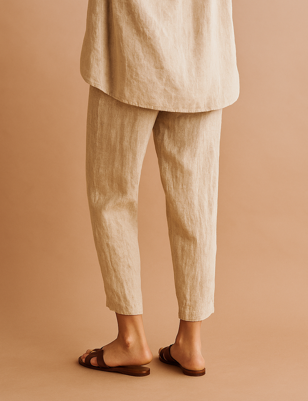 Tapered ankle-length linen pants in a natural beige tone with subtle vertical creasing, paired with flat brown leather sandals.