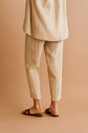 Tapered ankle-length linen pants in a natural beige tone with subtle vertical creasing, paired with flat brown leather sandals.
