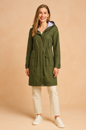 Olive green hooded raincoat with striped lining, front drawstring waist, flap pockets, and button details, styled with cream trousers and white sneakers.