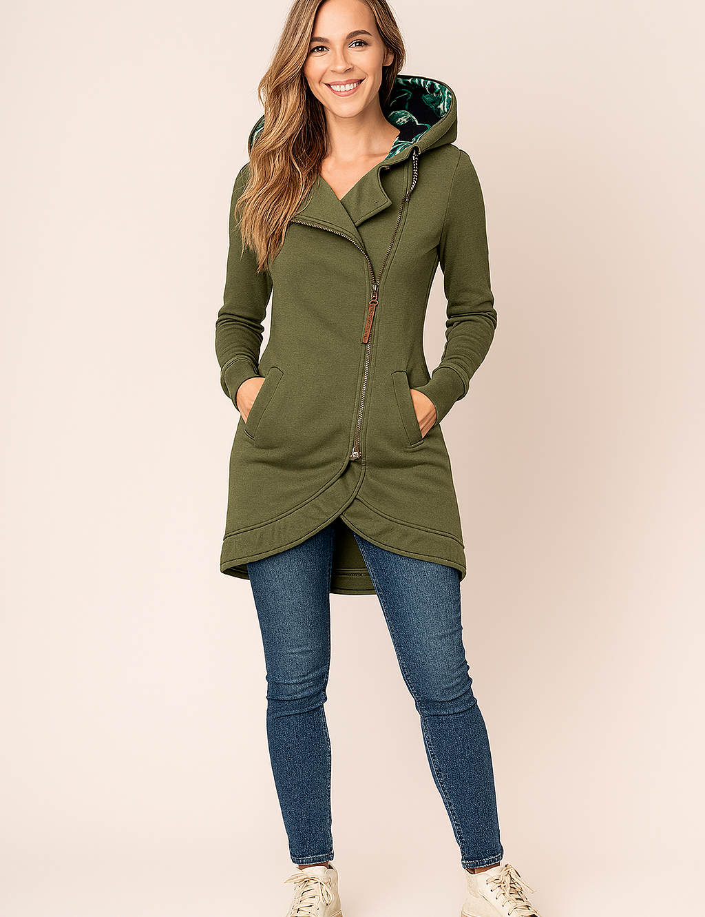 Olive green asymmetrical zip-up hoodie with curved hem, front pockets, and a tropical leaf-print hood lining, styled with slim blue jeans and cream high-top sneakers.