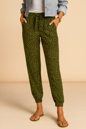 Olive green jogger pants with a black leopard print, elastic waistband and ankle cuffs, styled casually with a denim jacket and flat sandals.