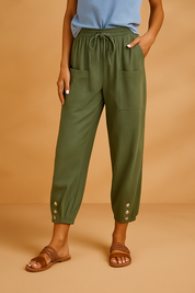 Olive green cropped linen pants with a drawstring waist, oversized front patch pockets, and silver button cuffs at the ankles, styled with woven brown sandals.