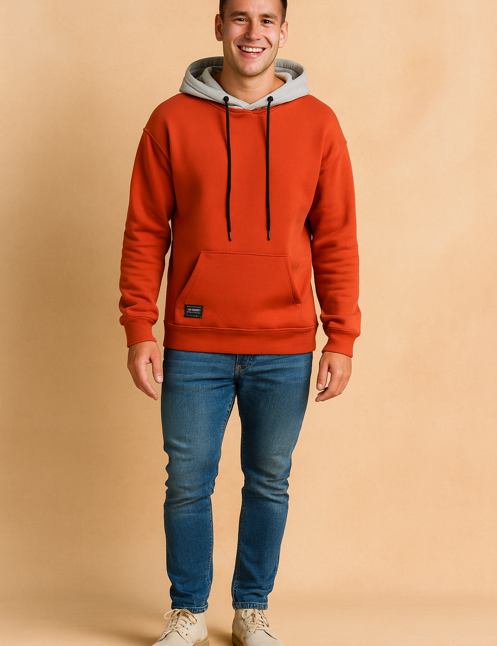 Men’s casual orange hoodie with grey hood lining, featuring black drawstrings and a front kangaroo pocket, styled with slim-fit blue jeans and light beige lace-up shoes.