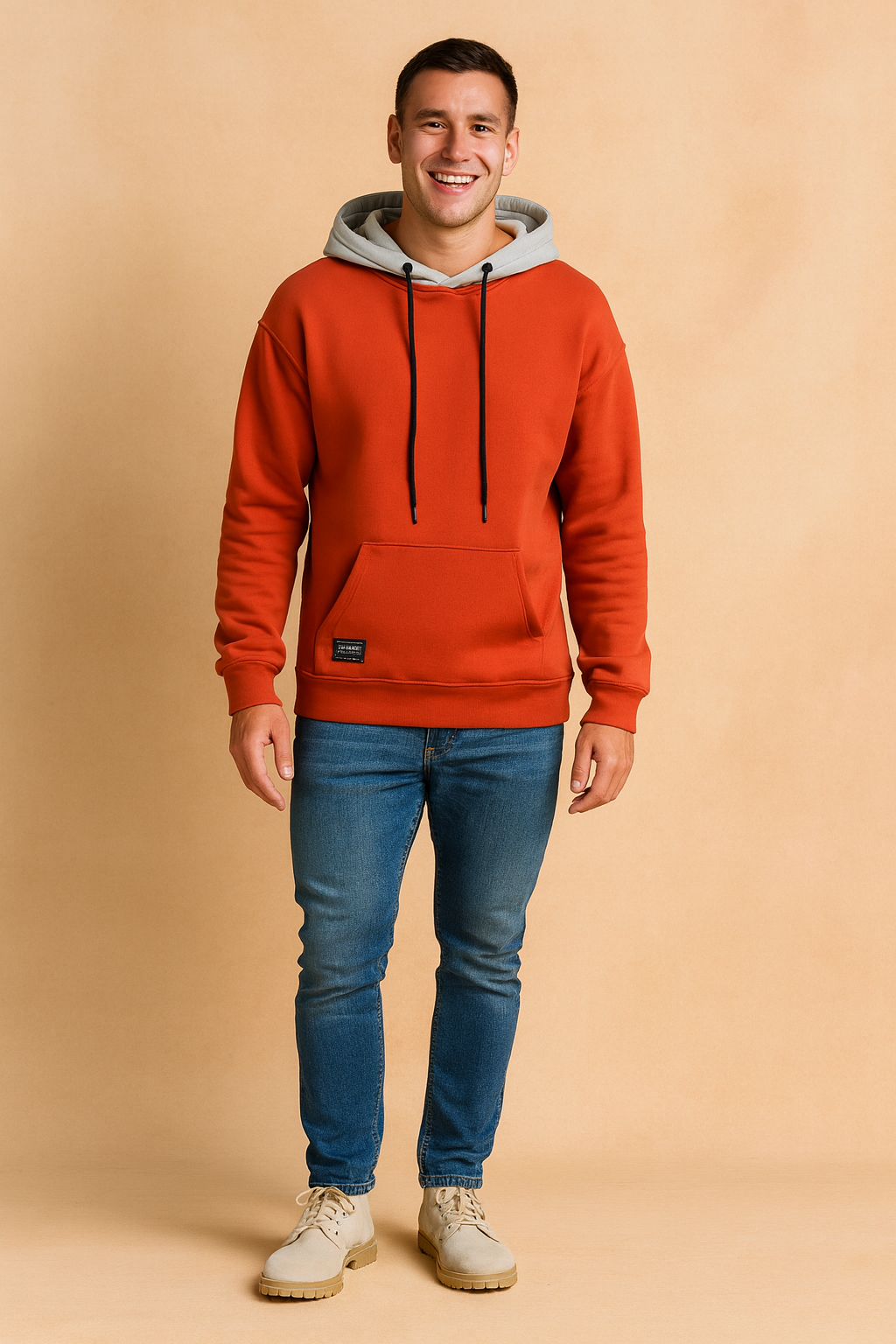 Men’s casual orange hoodie with grey hood lining, featuring black drawstrings and a front kangaroo pocket, styled with slim-fit blue jeans and light beige lace-up shoes.