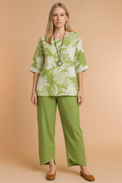 Two-piece sage green linen set featuring a short-sleeve top with oversized white floral print and matching wide-leg pants with asymmetrical hems, styled with a black pendant necklace and tan flats.