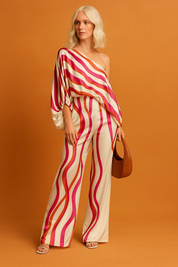 Cream satin two-piece set with magenta, red, and orange wavy stripes, designed with an off-shoulder oversized blouse and matching high-waisted wide-leg trousers, accessorized with a brown crescent bag and heeled sandals.