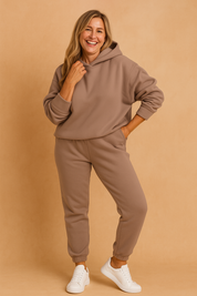 Taupe oversized hoodie and matching jogger pants in a soft fleece-textured fabric, styled casually with white sneakers.