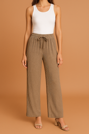 Taupe wide-leg pleated pants with a ribbed texture and adjustable drawstring waistband, styled with a fitted white tank top and nude block-heeled sandals.