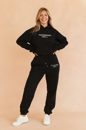 Two-piece black co-ord set featuring a relaxed-fit hoodie and high-waisted jogger pants with drawstring waist, both embellished with minimal white logo text for a clean streetwear look.
