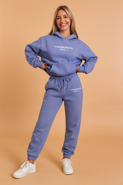 Oversized periwinkle hoodie and jogger set featuring drawstring waist, elastic cuffs, and “THATSSOFAITH SERIES 2” printed branding on both pieces, styled with classic white sneakers.