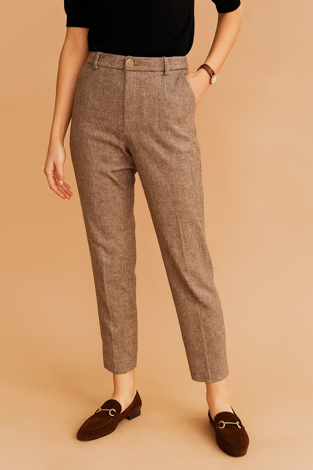Tailored taupe herringbone trousers with a classic front pleat, high waist, and ankle-length cut, paired with dark brown suede loafers featuring gold hardware.