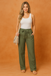 Olive green wide-leg linen drawstring pants paired with a white ribbed tank top, styled with beige sandals and a shoulder bag, photographed in a warm sand-colored studio.