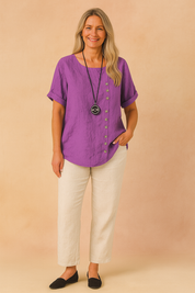Purple short-sleeve linen blouse with asymmetrical vertical button detail and round neckline, styled with off-white linen pants.