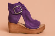 Purple suede wedge sandal with open toe, wide weathered ankle strap and oversized silver buckle, set on a natural wood-textured platform sole.