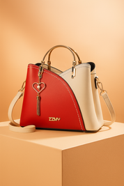 Red and ivory white structured faux leather handbag with gold-tone handles, heart-shaped pearl tassel charm, and contrast stitching, placed on a warm beige platform in soft luxury lighting.
