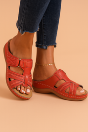 Red leather slip-on sandals with wide crisscross straps, intricate perforated detailing, secure Velcro closure, and contrast stitching on a cushioned wedge sole.