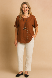 Rust brown short-sleeve linen blouse with round neckline and asymmetric vertical button detail, styled with off-white straight-leg pants.