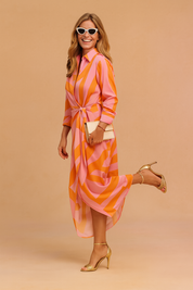 Long-sleeve midi dress featuring a vivid geometric swirl print in bold pink and orange tones, with a wrap-tied waist, shirt collar, high-low hemline, and paired with gold high-heeled sandals.
