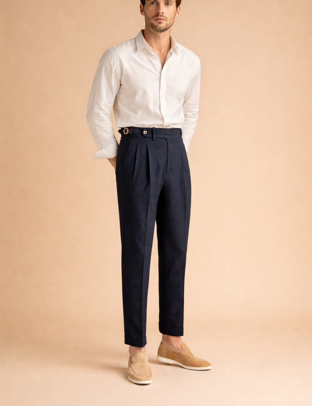 Tailored high-waist pleated trousers with a structured waistband, front button closure, belt side adjusters, clean tapered leg, and cuffed hem styled with a long-sleeve button-up shirt and minimalist loafers.