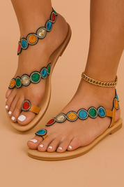 Tan flat sandals adorned with embroidered circular patches in vibrant red, orange, green, blue, pink, and silver, each outlined in black and gold stitching, featuring a toe loop and ankle strap for a secure fit.