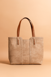Textured beige tote bag with rich brown handles, featuring detailed stitching and vertical seam accents, captured in a softly lit studio setup with a warm sand and pastel blush backdrop.
