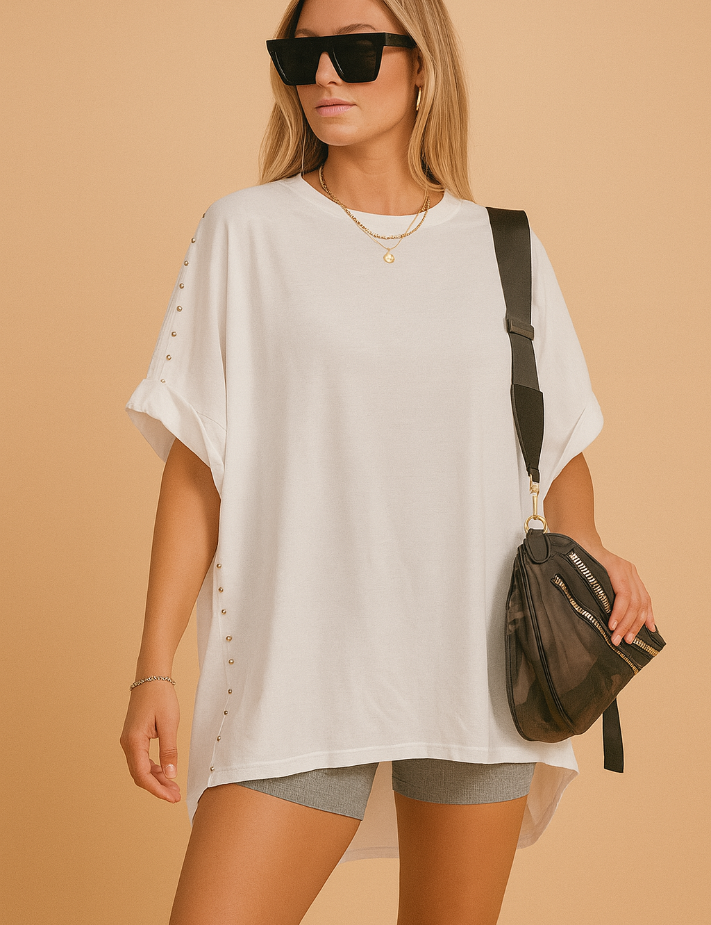 Oversized white short-sleeve poncho T-shirt with silver stud detailing down the sides, paired with mid-rise gray biker shorts and accessorized with a black camo crossbody bag featuring gold zippers.