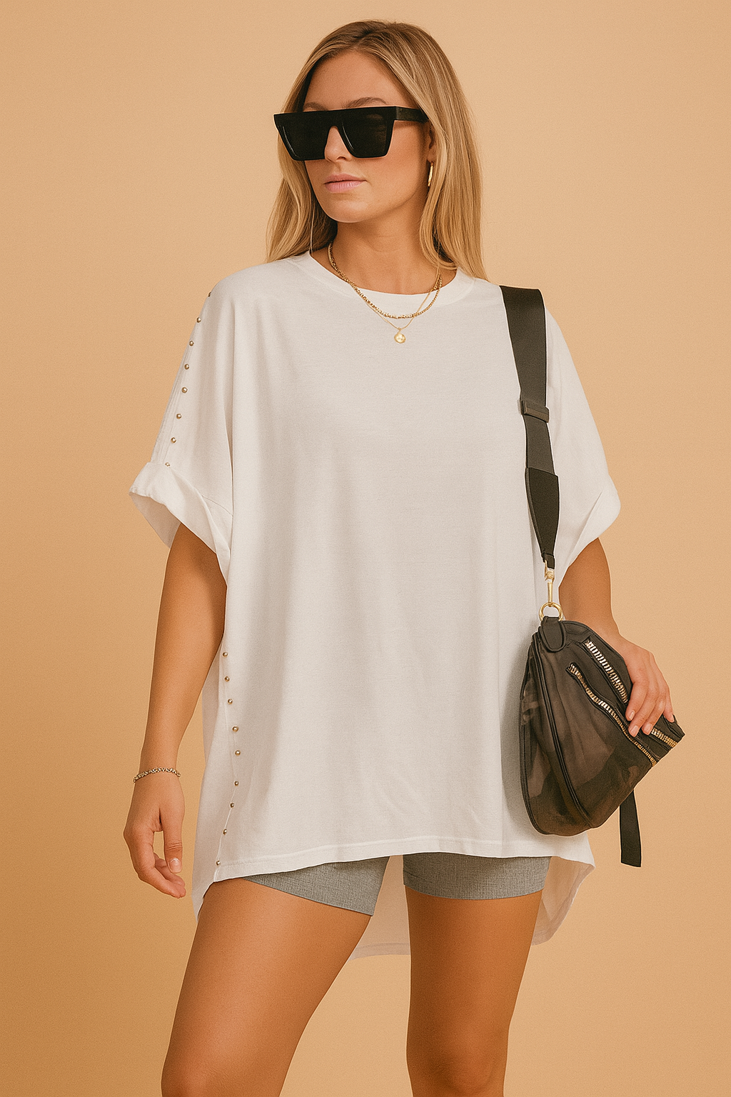 Oversized white short-sleeve poncho T-shirt with silver stud detailing down the sides, paired with mid-rise gray biker shorts and accessorized with a black camo crossbody bag featuring gold zippers.