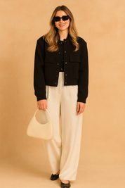Black cropped bomber jacket with snap buttons and front pockets, paired with wide-leg off-white trousers, black shoes, sunglasses, and a cream handbag.