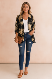 Czarny damski kardigan kimono w kwiaty z kremowo-beżowym nadrukiem róż, z drapowanym przodem i krótkimi rękawami, noszony na białej bluzce z dekoltem w serek, obcisłych niebieskich jeansach z przetarciami, brązowych sandałach na klockowym obcasie, warstwowych złotych naszyjnikach, bransoletce i brązowej kopertówce.
