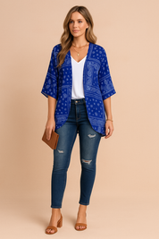 Damski kardigan kimono w kolorze niebieskim z nadrukiem bandany, z krótkimi rękawami i drapowanym przodem, noszony na białej bluzce z dekoltem w serek, obcisłych niebieskich jeansach z przetarciami, brązowych sandałach na klockowym obcasie, warstwowych złotych naszyjnikach, bransoletce i brązowej kopertówce.