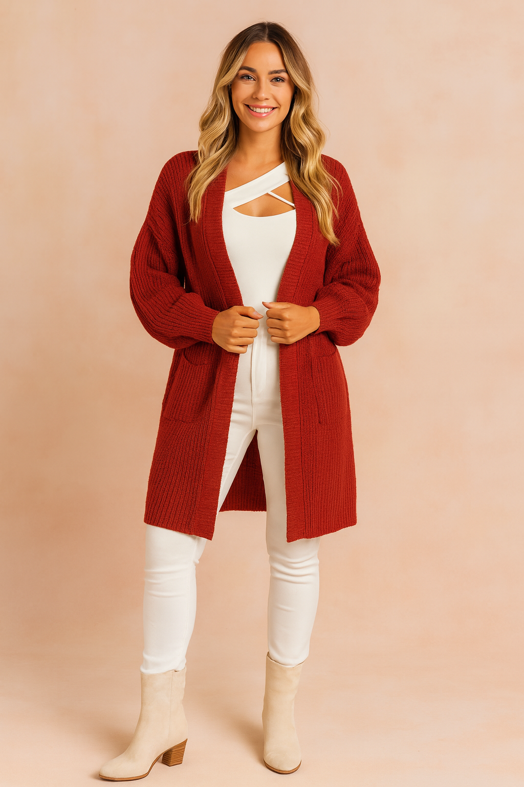 Deep red oversized chunky knit cardigan with open front, long sleeves, and two front patch pockets, styled over a white top and white fitted pants with beige suede ankle boots.