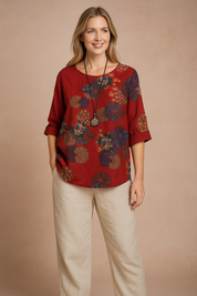 Boho-inspired red blouse with vibrant multicolor circular floral patterns, diagonal button accents at the neckline, and mid-length sleeves, styled over loose beige trousers for a casual artistic look.