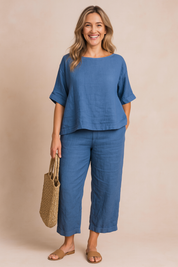 Two-piece blue linen set featuring a relaxed-fit elbow-sleeve top and cropped wide-leg pants with subtle texture, styled with tan slide sandals and a woven tote bag.