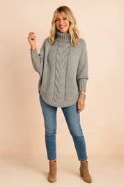 Oversized grey cable-knit turtleneck sweater with long sleeves, paired with medium-wash distressed skinny jeans and taupe suede ankle boots, accessorized with silver bangles.