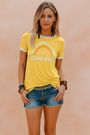 Retro yellow "GOOD VIBES" graphic T-shirt with a faded rainbow print, styled with denim shorts and layered boho accessories, perfect for casual summer fashion.