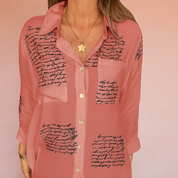 Flowy rose-pink button-down blouse with black handwritten script print on sheer fabric, featuring a relaxed collar, front buttons, and chest patch pocket detail.