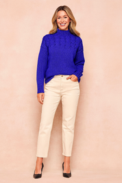 Royal blue high-neck cable-knit sweater with intricate braided detailing on the upper chest and shoulders, paired with cream straight-leg trousers and black pointed-toe heels.