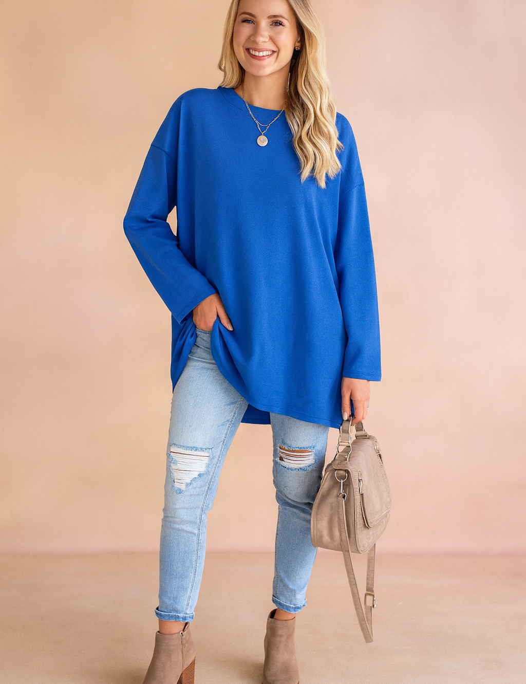 Royal blue oversized long-sleeve tunic paired with light-wash distressed skinny jeans, taupe suede ankle boots, and a matching beige handbag.