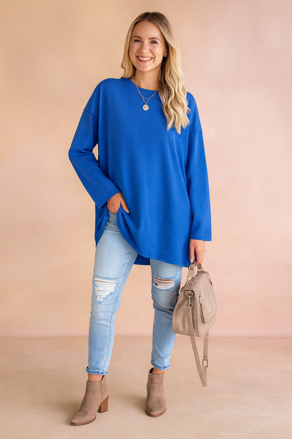 Royal blue oversized long-sleeve tunic paired with light-wash distressed skinny jeans, taupe suede ankle boots, and a matching beige handbag.