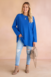 Royal blue oversized long-sleeve tunic paired with light-wash distressed skinny jeans, taupe suede ankle boots, and a matching beige handbag.