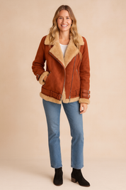 Rust-red shearling aviator jacket with beige faux shearling lining, detailed with zip closure, buckled straps, and hem belt, styled with light blue straight-leg jeans and black ankle boots
