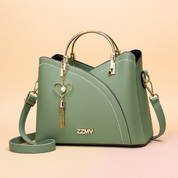 Sage green structured handbag with black trim and white stitching, gold-tone hardware, heart charm with pearl and tassel, and matching shoulder strap, placed on a soft gradient beige-pink background.