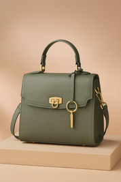 Structured sage green leather handbag with top handle, gold turn-lock clasp, key charm detail, and detachable shoulder strap, displayed on a sand-toned platform.