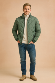 Sage green quilted jacket with V-shaped stitching, zip closure, black-trimmed zipper pockets, styled with a white turtleneck, slim blue jeans, and beige suede boots.