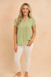 Sage green short-sleeve blouse with navy heart prints and soft pleated front detail, styled with slim-fit white pants and white sandals.