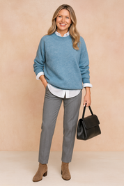 Sky-blue oversized sweater layered over a white button-up shirt, paired with gray tailored trousers, taupe ankle boots, and a black leather handbag.