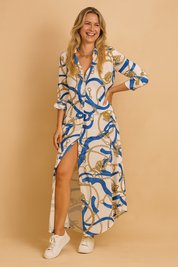 Long-sleeve shirt dress with a vivid blue and gold baroque-style chain print on a white base, featuring a front button-down design, asymmetric hemline, and high side slit, paired with white sneakers for a chic yet relaxed look.