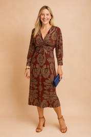 Elegant burgundy midi dress with intricate gold paisley print, long sleeves, ruched front detail, and a modest V-neckline—styled with tan heels and a royal blue clutch for a polished, sophisticated look.