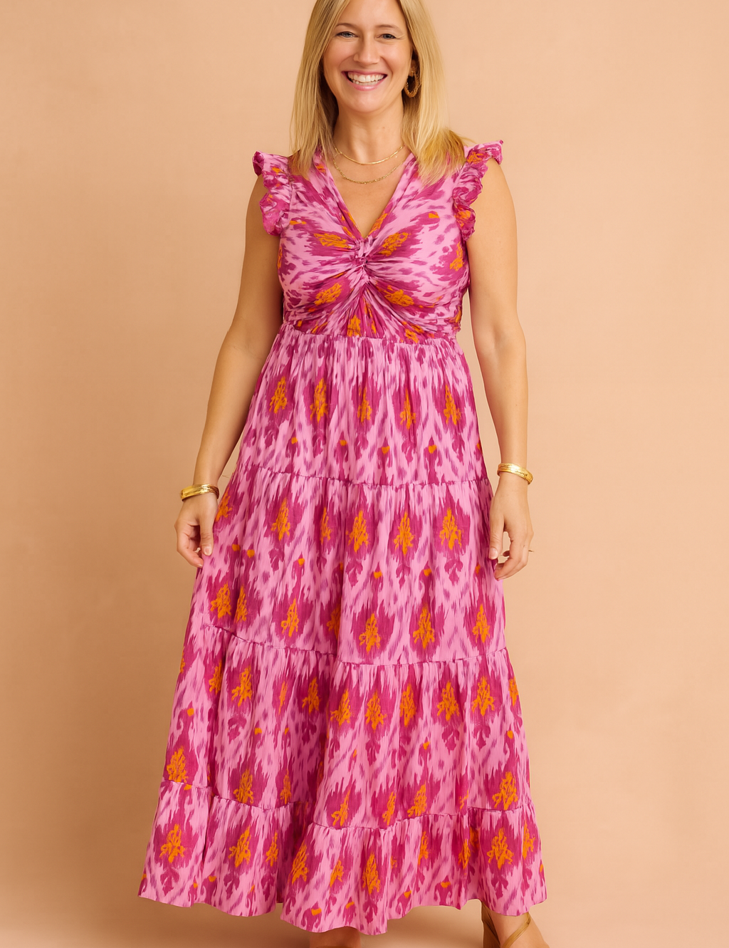 Sleeveless pink maxi dress with a vibrant ikat print in purple and orange tones, featuring a ruched twist-front bodice, ruffled shoulders, and a tiered full-length skirt.