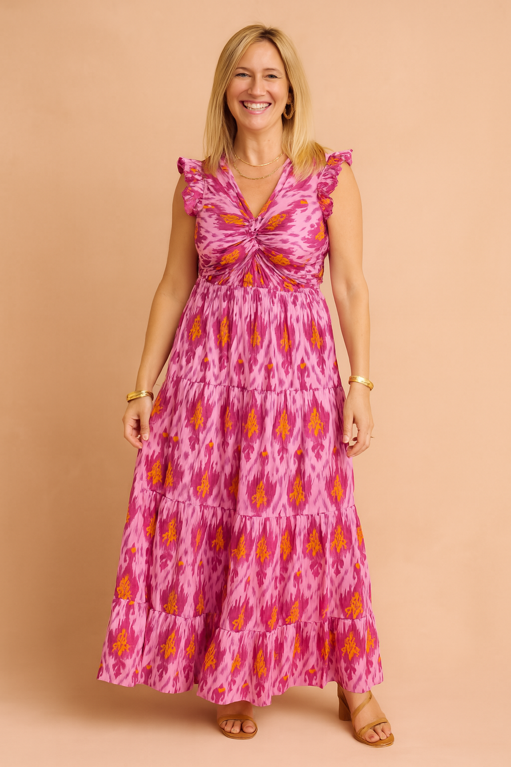 Sleeveless pink maxi dress with a vibrant ikat print in purple and orange tones, featuring a ruched twist-front bodice, ruffled shoulders, and a tiered full-length skirt.